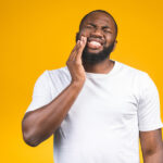 Black man in a white t-shirt against a yellow background cringes in pain and touches his cheek due to a dental abscess