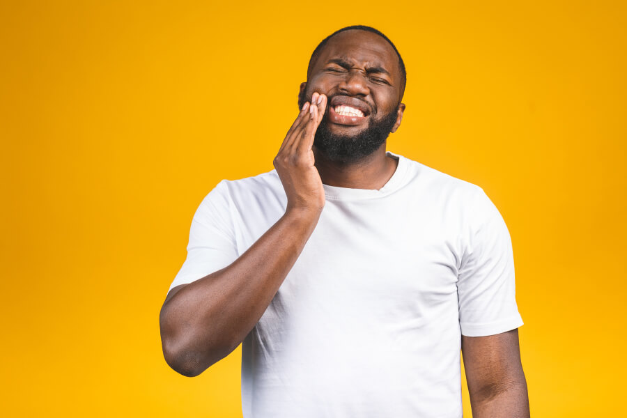 Black man in a white t-shirt against a yellow background cringes in pain and touches his cheek due to a dental abscess